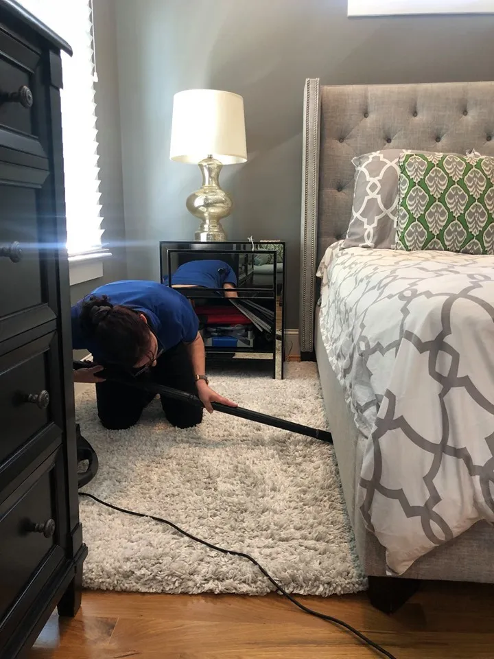 An image of a lady cleaning the underside of the bed with a vacuum.